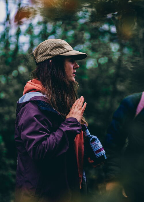 Eine Frau mit brauner Baseballkappe und lila Jacke steht in einem Wald und hält eine Wasserflasche in der Hand.