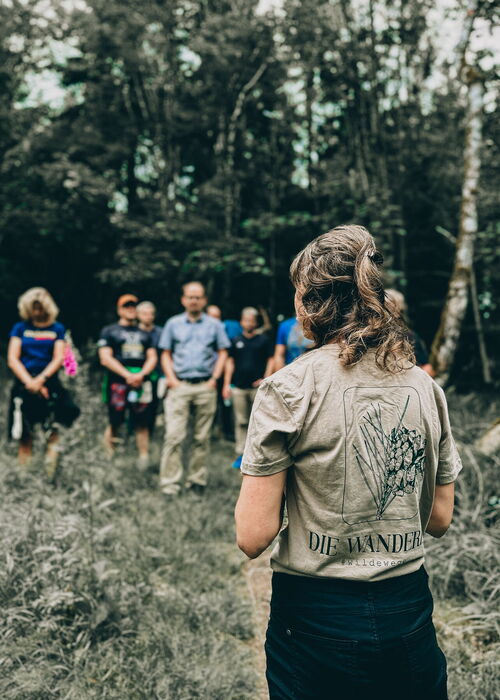 Eine Gruppe von Menschen steht auf einem Pfad in einem Wald, während eine Person im Vordergrund mit dem Rücken zur Kamera steht und ein T-Shirt mit der Aufschrift „DIE WANDERER“